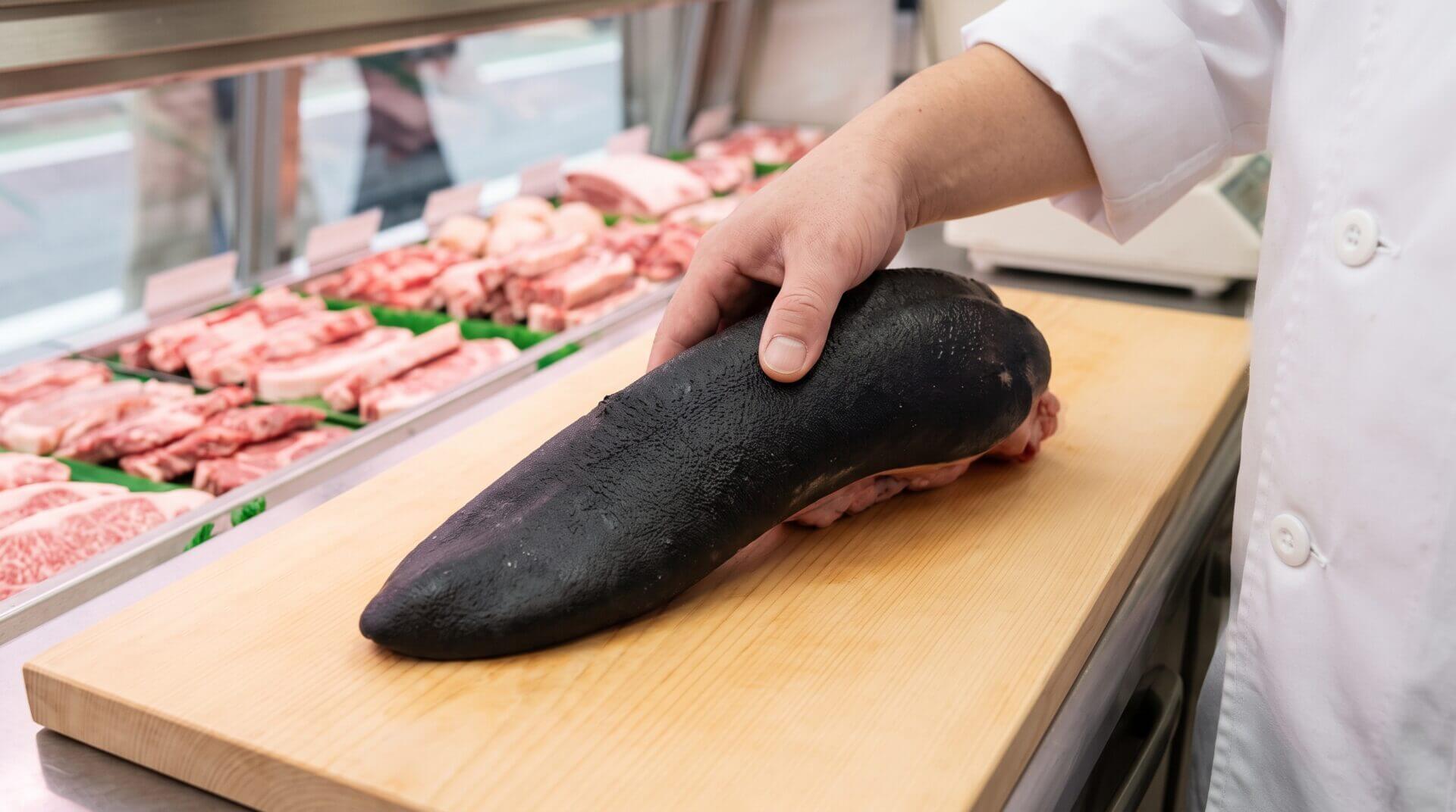 日本の精肉店で、白い作業着を着た日本人の職人が、黒毛和牛の特徴である黒い外皮に覆われた生の牛タン原木（黒タン）を手に持ち、木製のまな板の上で確認している様子。自然光で rough な黒い外皮の質感が強調されている。