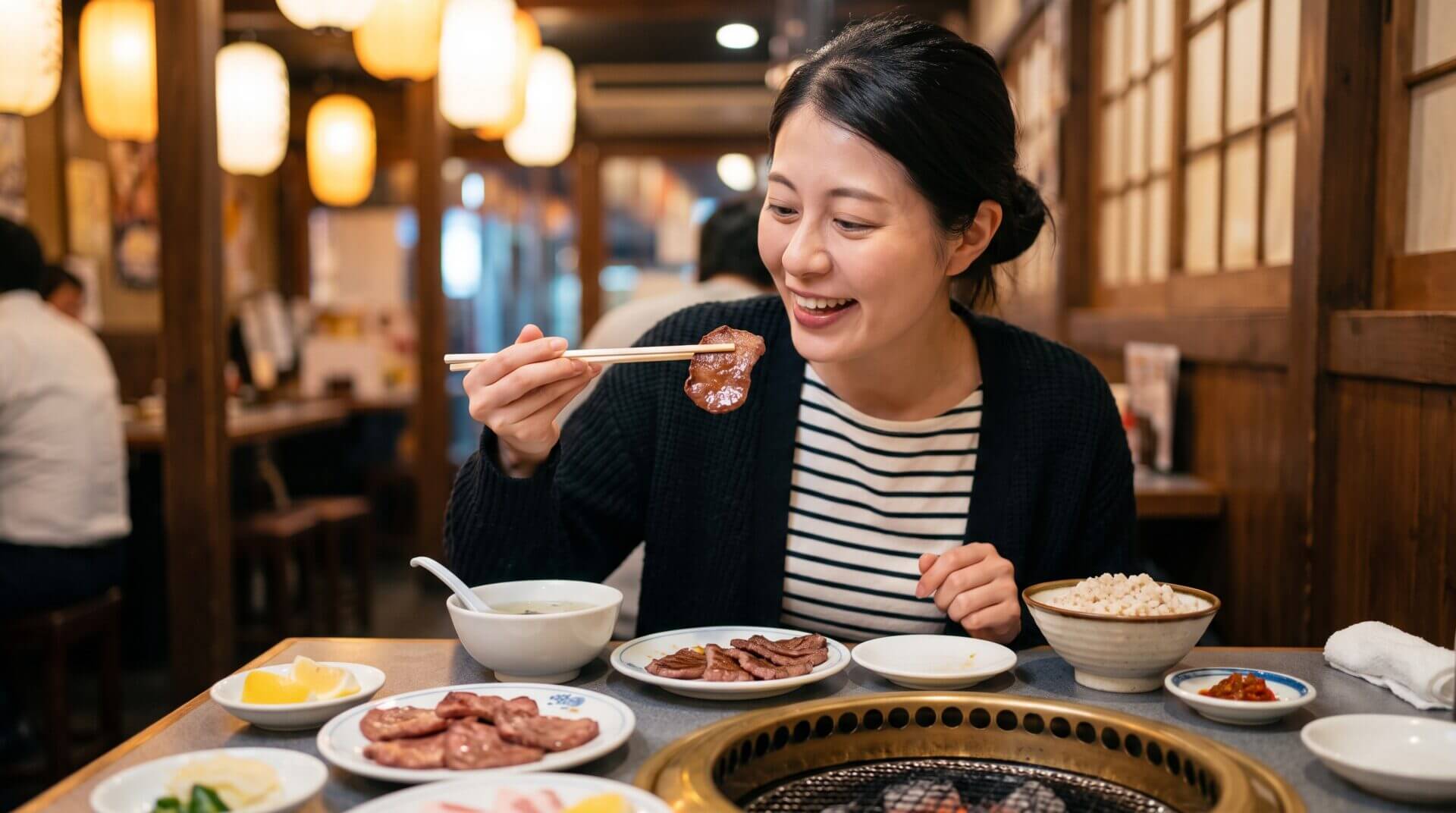 提灯が飾られた賑やかな日本の焼肉店で、日本人の女性が笑顔で牛タンを食べている。テーブルには七輪や麦飯、スープが並ぶ。