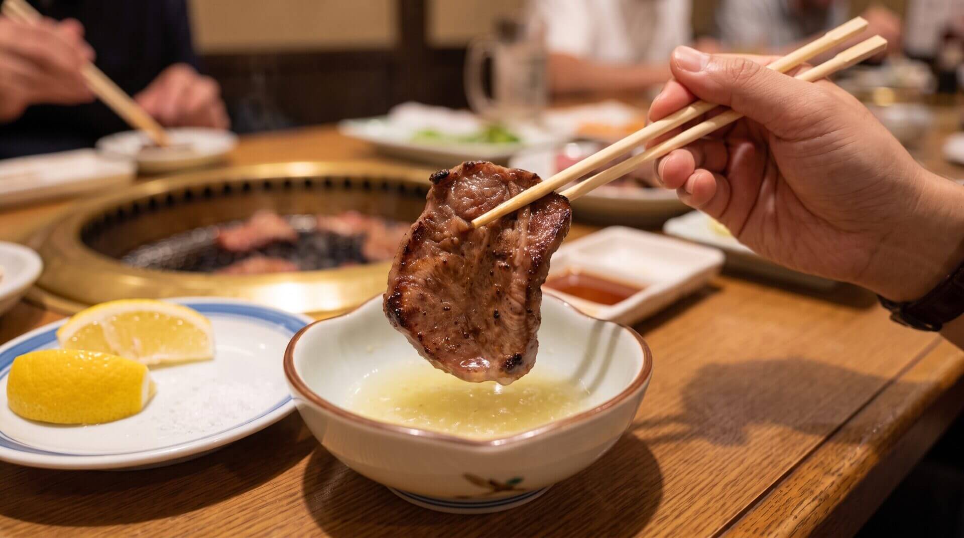 焼肉店で箸を使い、焼きたての牛タンを小鉢に入ったレモンダレに浸す瞬間の手元のアップ。